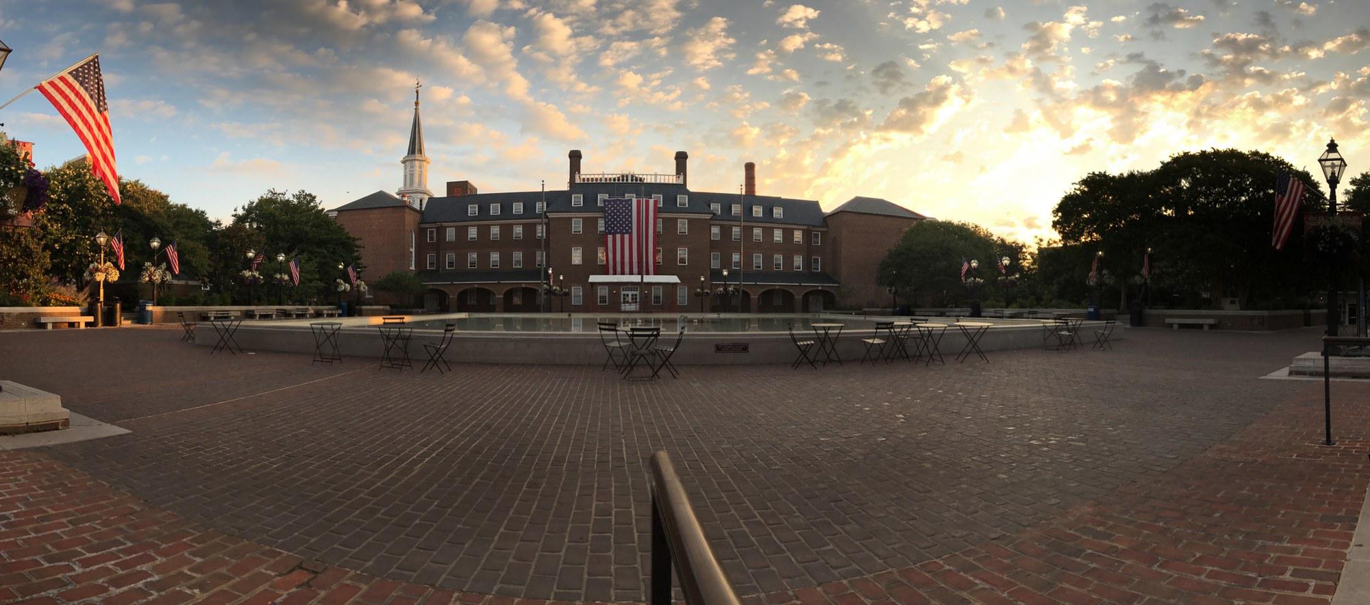 Wide panoramic photograph at sunrise of Market Square in front of Alexandria City Hall: brick-paved plaza in the foreground with bistro tables ringing a low oval fountain pool; the Federal-style brick city hall building behind, with a large American flag hanging from the central facade and the clock tower spire visible at left; rows of American flags on the surrounding lamp posts; high cumulus clouds catching warm sunrise light over the rooftops.