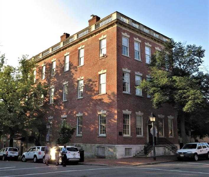 Color photograph of the Bank of Alexandria, a three-story Federal-style red-brick building at the corner of North Fairfax and Cameron Streets in Old Town Alexandria. The corner facade is framed by mature trees; a balustrade with white spindles runs across the roof line; the building's stone base, dressed-stone window heads, and tall double-hung sash windows are visible. Modern cars and pedestrians on the street at right.