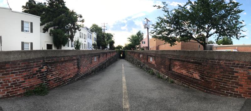 Wide panoramic photograph from the western approach to the Wilkes Street Tunnel in Old Town Alexandria. The asphalt path descends into a short brick-vaulted railroad tunnel framed by stone-and-brick retaining walls on each side, with the western portal visible at center-distance as a small dark arch. White-painted Federal-style row houses line the left bank of the cut and a brick warehouse and mature tree line the right; high cumulus clouds catch warm morning light over the rooftops.