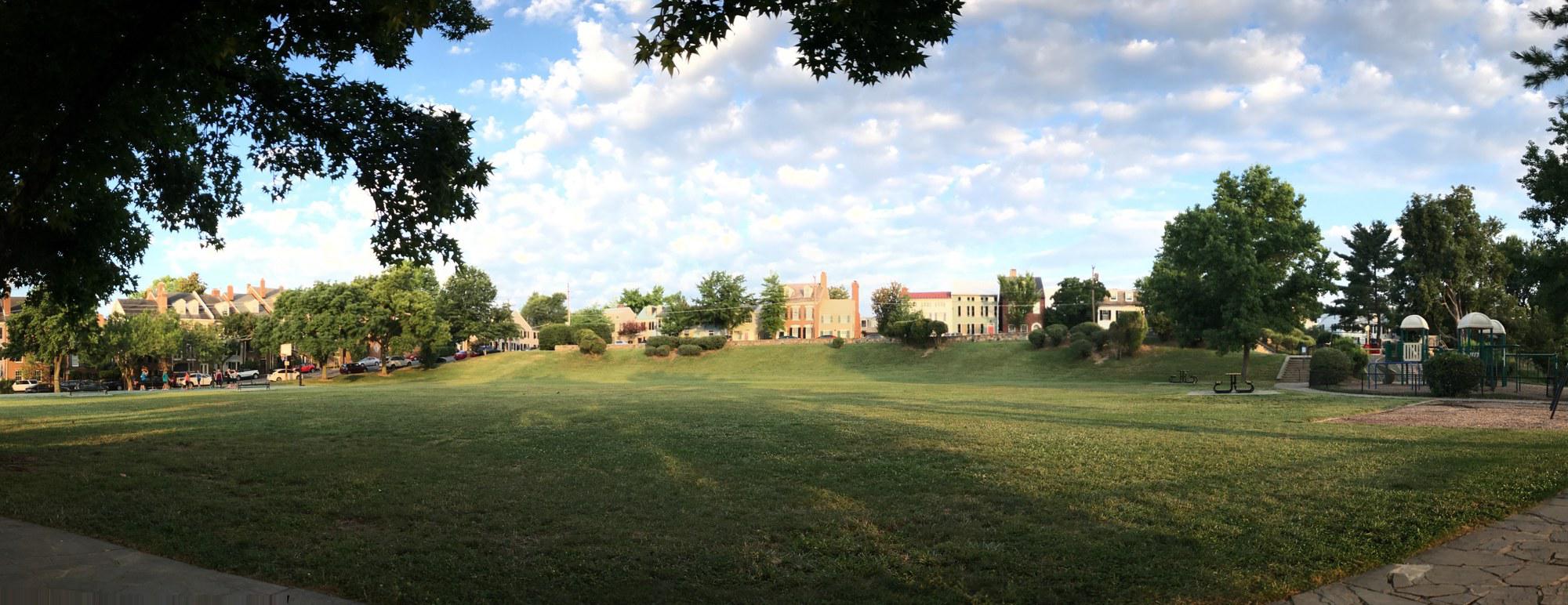 Wide panoramic photograph at sunrise of Windmill Hill Park at 501 South Union Street in Old Town Alexandria. Broad open grass meadow in the foreground sloping gently down to the camera; the Old Town row houses on Union Street rise on the far side of the field; mature shade trees frame the panorama at left and right; a playground structure is visible at right; high cumulus clouds catch warm sunrise light over the rooftops.