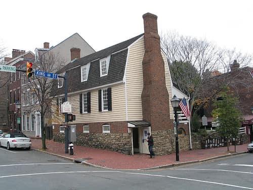Color photograph of the Ramsay House as the modern Alexandria Visitor Center — a small frame building with red gambrel roof and three dormer windows at the corner of King and Fairfax Streets, with the brick chimney prominent on the gable end. Stone retaining wall and brick walkway in foreground; a blue "KING ST" street sign at left; modern lampposts; American flag visible at right.
